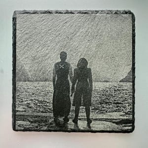 Puede incluir: Una foto en blanco y negro de dos personas de pie en una playa, mirando hacia el agua. La foto está grabada en una baldosa de pizarra negra.