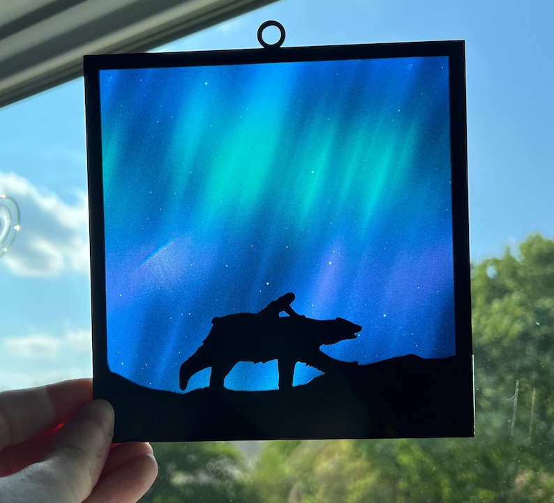May include: A black silhouette of a polar bear walking under a vibrant green and blue aurora borealis. The aurora is painted on a square piece of glass with a hanger at the top.