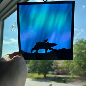 May include: A stained glass window hanging with a black silhouette of a bear and a person walking in front of a blue and green aurora borealis.