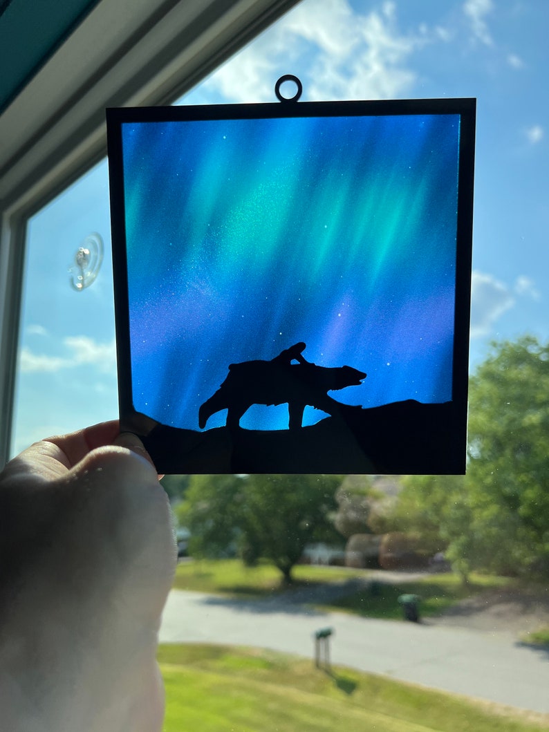 May include: A stained glass window hanging with a black silhouette of a bear and a person walking in front of a blue and green aurora borealis.