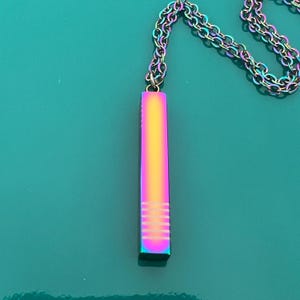 Puede incluir: Un collar con colgante vertical con acabado iridiscente arcoíris. El colgante es rectangular con líneas horizontales grabadas cerca de la parte inferior. La cadena es de un color iridiscente a juego. El collar se muestra sobre un fondo turquesa.