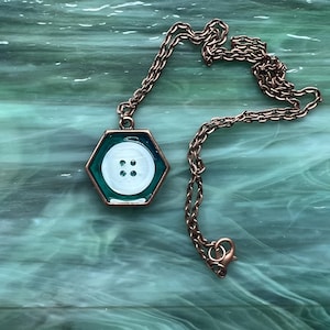 May include: A copper-colored chain necklace with a hexagonal pendant. The pendant features a white button with four holes set against a teal background. The necklace has a lobster clasp.