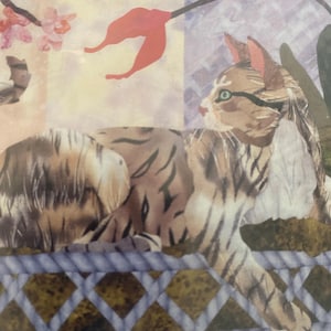May include: A detailed illustration of a cat with brown and black stripes, resting on a blue woven surface. The cat has green eyes and is set against a backdrop of floral elements and a patterned background.