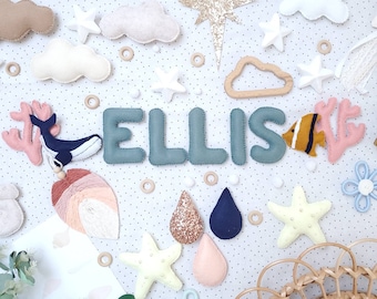 May include: A whimsical nursery decor arrangement featuring the name "ELLIS" in teal felt letters. The scene includes felt clouds, stars, a whale, coral, a fish, and decorative elements. The background is a white surface with small black polka dots.