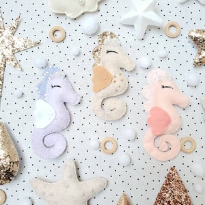 May include: Three felt seahorse ornaments with glitter accents. The seahorses are pink, purple, and beige. The ornaments are surrounded by white pom-poms, wooden rings, and felt stars.