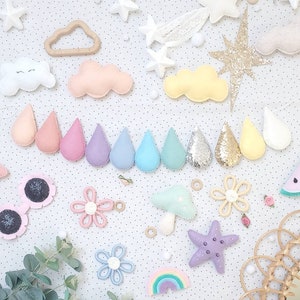 May include: A collection of felt craft decorations in various shapes and colors, including clouds, stars, raindrops, flowers, a mushroom, a rainbow, and a starfish. The decorations are arranged on a white background with small white dots.
