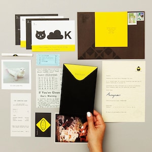 May include: A black envelope with a yellow flap labeled "Puzzles" is open to reveal a series of puzzles. The envelope is on a table with other clues, including a newspaper clipping, a photo of a seal, and a postcard with a photo of a building.