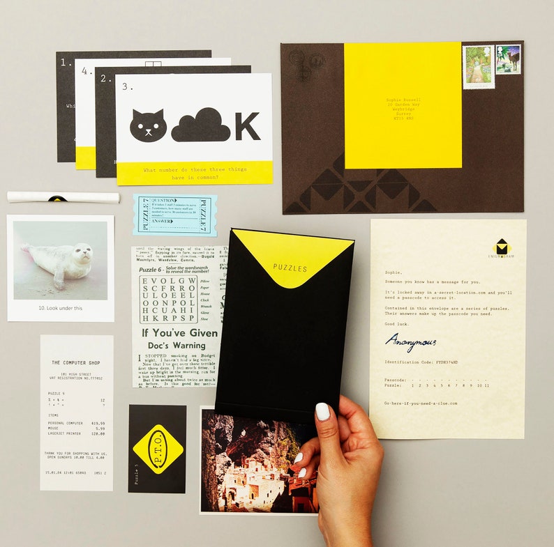 May include: A black envelope with a yellow flap labeled "Puzzles" is open to reveal a series of puzzles. The envelope is on a table with other clues, including a newspaper clipping, a photo of a seal, and a postcard with a photo of a building.