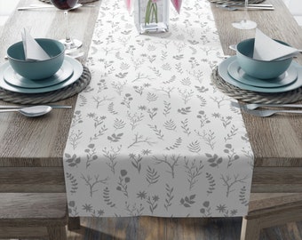 May include: A white table runner with a gray floral pattern, set on a wooden table. The table is set with teal-colored bowls, plates, and wine glasses. A vase of pink lilies sits in the center.