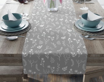 May include: A gray table runner with a white floral pattern, set on a wooden table. The runner features various botanical designs, including leaves and branches. The table is set with plates, bowls, and wine glasses.
