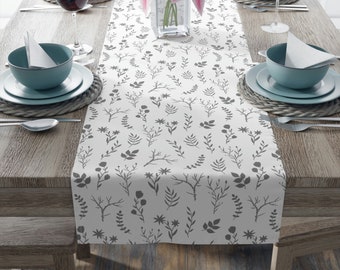 May include: A white table runner with a gray floral pattern. The runner is laid out on a wooden table with place settings on either side.