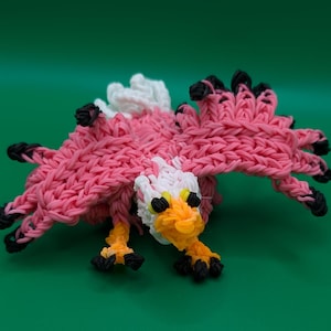 May include: A pink rubber band eagle with black eyes, an orange beak, and white feathers on its head. The eagle is made with a loom and is a popular craft project.