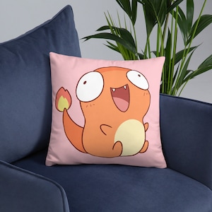 May include: A pink pillow with a cartoon illustration of a smiling orange creature with a flame tail. The creature has large eyes and a wide open mouth.
