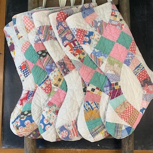May include: Five quilted Christmas stockings made from assorted vintage fabric scraps in various colors and patterns. Each stocking features a white fabric cuff and a fabric loop for hanging, showcasing a patchwork design with stitched textures.