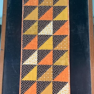May include: A rectangular quilted table runner featuring a geometric pattern of half-square triangles. The fabric blocks alternate between black with a candy corn print, solid mustard yellow, orange with a subtle pattern, and cream floral prints.
