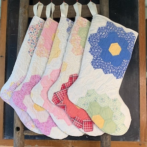 May include: Five quilted Christmas stockings featuring a patchwork hexagon pattern. Each stocking displays a unique combination of floral, plaid, and solid fabric prints in shades of blue, pink, yellow, red, and green, finished with a white fabric loop.