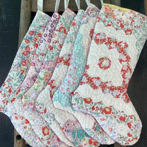 May include: Six quilted Christmas stockings made from various floral patterned fabrics in shades of red, pink, blue, and green. Each stocking features a white fabric hanging loop at the top and intricate stitching throughout the quilted surface.
