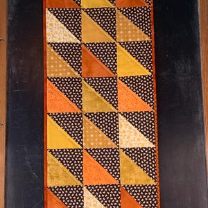 May include: A rectangular quilted table runner featuring a geometric pattern of half-square triangles. The design alternates between black fabric with a small candy corn print and various solid or patterned fabrics in shades of orange, yellow, and cream.