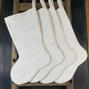May include: A set of four off-white quilted stockings, each featuring a textured, stitched pattern and a fabric hanging loop at the top. The stockings are uniform in color and design, displaying a soft, crinkled fabric surface throughout.