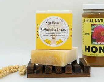 Puede incluir: Una barra de jabón amarilla con una etiqueta blanca que dice "Kay Pedals Oatmeal & Honey Natural Crafted, For Gentle Skin Soap". El jabón está sobre un jabonera de madera junto a un frasco de vidrio transparente de miel etiquetado como "Local Natural Raw Wildflower Honey 100% Net Weight 16 oz (1 lb) 454 g".