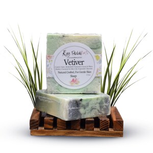 Peut inclure: Deux savons au vétiver vert et blanc avec une étiquette indiquant "Kay Pedals" et "Vetiver" sur un porte-savon en bois. Le savon est fabriqué naturellement pour les peaux sensibles.