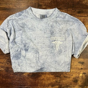 May include: A light blue t-shirt with a faded white print of a medical symbol and the text "Emergency Department".