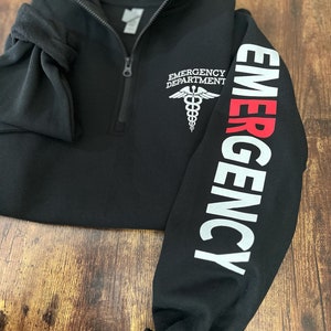 May include: Black fleece pullover with a zipper and a white embroidered medical symbol on the left chest. The right sleeve has the word "EMERGENCY" printed in white with the first letter in red.