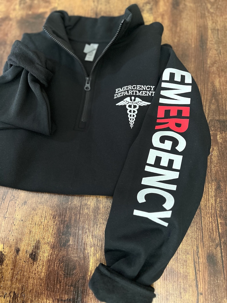 May include: Black fleece pullover with a zipper and a white embroidered medical symbol on the left chest. The right sleeve has the word "EMERGENCY" printed in white with the first letter in red.