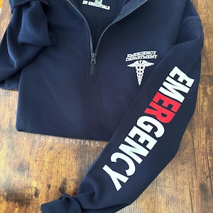 May include: Navy blue 1/4 zip sweatshirt with the text "ER Essentials" on the front and the text "EMERGENCY" on the sleeve in white with a red "E". The back of the sweatshirt has a white medical symbol with the text "EMERGENCY DEPARTMENT" above it.
