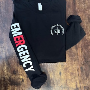 May include: Black long-sleeve shirt with the word "EMERGENCY" in white and red lettering on the left sleeve. The right sleeve is rolled up. The shirt has "EMERGENCY DEPARTMENT ED" printed on the chest.