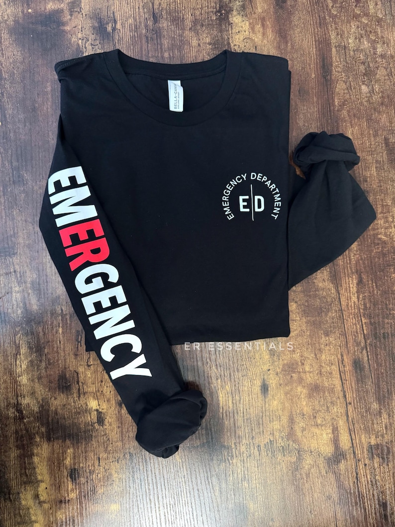 K&ouml;nnte beinhalten: Schwarzes Langarmshirt mit dem Wort "EMERGENCY" in wei&szlig;er und roter Schrift auf dem linken &Auml;rmel. Der rechte &Auml;rmel ist hochgekrempelt. Das Shirt hat "EMERGENCY DEPARTMENT ED" auf der Brust aufgedruckt.