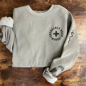 May include: A gray sweatshirt with the text "Emergency Room Nurse" in a circle with a plus sign in the center. The right sleeve has a caduceus symbol.