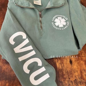 May include: A sage green sweatshirt with a white zipper and a white graphic on the front that says "Cardiovascular Intensive Care Unit" with a star of life symbol. The left sleeve has a white graphic that says "CVICU". The sweatshirt has the text "ER Essentials" on the bottom right.