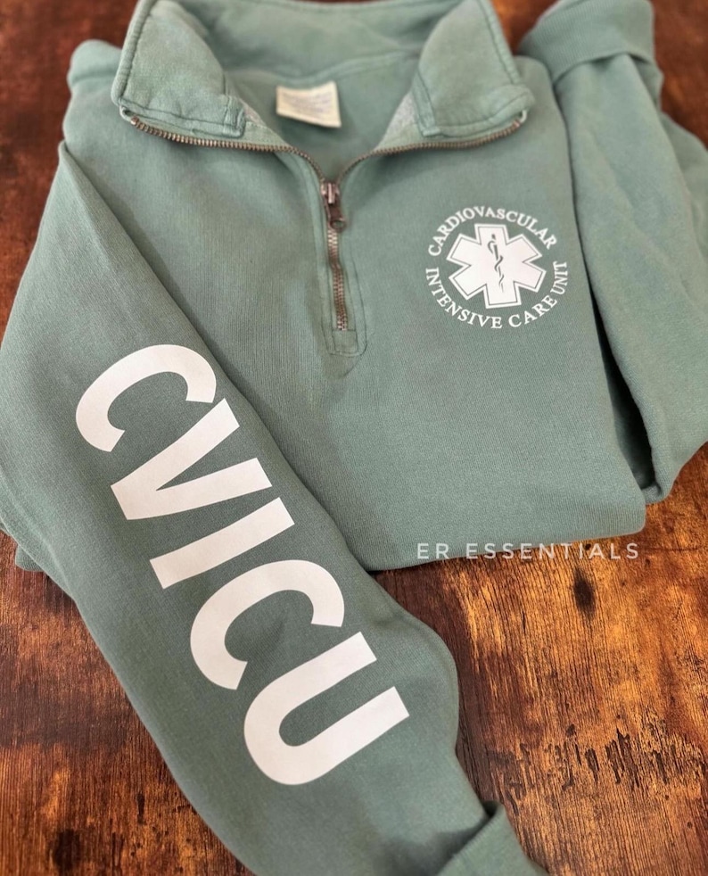 May include: A sage green sweatshirt with a white zipper and a white graphic on the front that says "Cardiovascular Intensive Care Unit" with a star of life symbol. The left sleeve has a white graphic that says "CVICU". The sweatshirt has the text "ER Essentials" on the bottom right.
