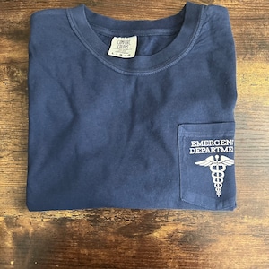 May include: A navy blue short-sleeve t-shirt with a crew neckline and a chest pocket. The pocket features a white graphic of a caduceus symbol and the text EMERGENCY DEPARTMENT. The shirt has a Comfort Colors brand label at the inner neck.