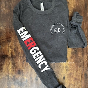 K&ouml;nnte beinhalten: Dunkelgraues Langarmshirt mit dem Wort "EMERGENCY" in wei&szlig;er und roter Schrift auf dem linken &Auml;rmel. Das Shirt hat auch den Text "EMERGENCY DEPARTMENT ED" in einem kreisf&ouml;rmigen Design auf der Brust.