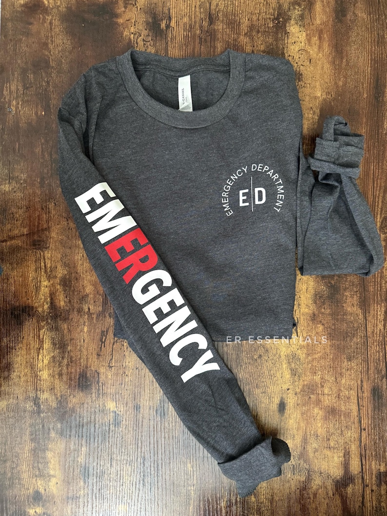 K&ouml;nnte beinhalten: Dunkelgraues Langarmshirt mit dem Wort "EMERGENCY" in wei&szlig;er und roter Schrift auf dem linken &Auml;rmel. Das Shirt hat auch den Text "EMERGENCY DEPARTMENT ED" in einem kreisf&ouml;rmigen Design auf der Brust.