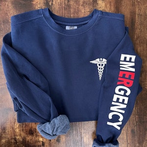 May include: Navy blue sweatshirt with a white medical symbol on the left chest and the word "EMERGENCY" in white and red on the right sleeve.