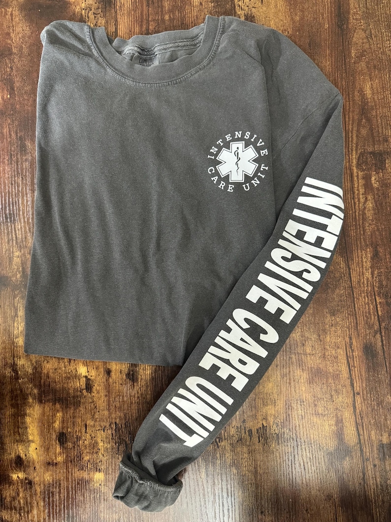 May include: A dark gray long-sleeve shirt with the words "INTENSIVE CARE UNIT" printed in white on the sleeve. The shirt features a white medical symbol and the words "INTENSIVE CARE UNIT" on the chest.