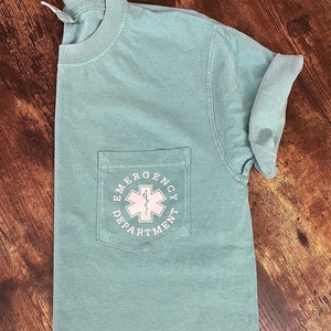 May include: A light teal t-shirt with a pocket featuring the text "EMERGENCY DEPARTMENT" surrounding a white medical symbol. The shirt has short sleeves and a crew neck.