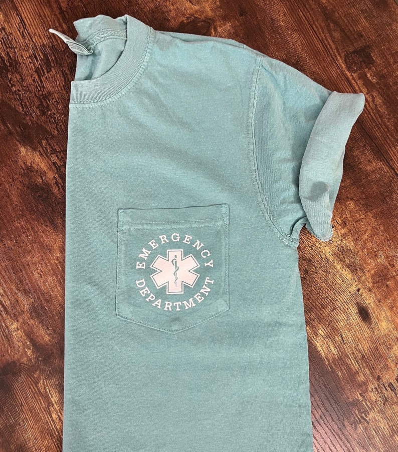 May include: A light teal t-shirt with a pocket featuring the text "EMERGENCY DEPARTMENT" surrounding a white medical symbol. The shirt has short sleeves and a crew neck.