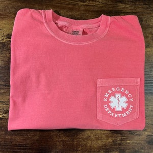 May include: Coral-colored t-shirt with a pocket featuring a white graphic of a medical symbol and the words "Emergency Department". The shirt is folded on a wooden surface.