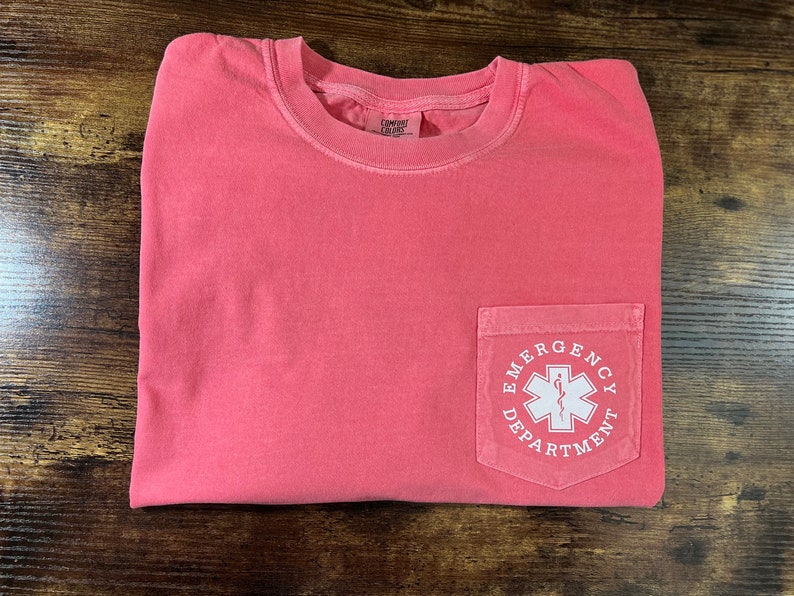 May include: Coral-colored t-shirt with a pocket featuring a white graphic of a medical symbol and the words "Emergency Department". The shirt is folded on a wooden surface.