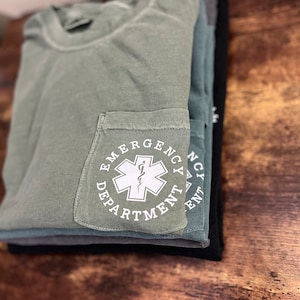 May include: A stack of folded t-shirts in shades of green, gray, and black. The top shirt is olive green with a pocket featuring the words "Emergency Department" around a white medical symbol. The shirts are neatly arranged on a wooden surface.