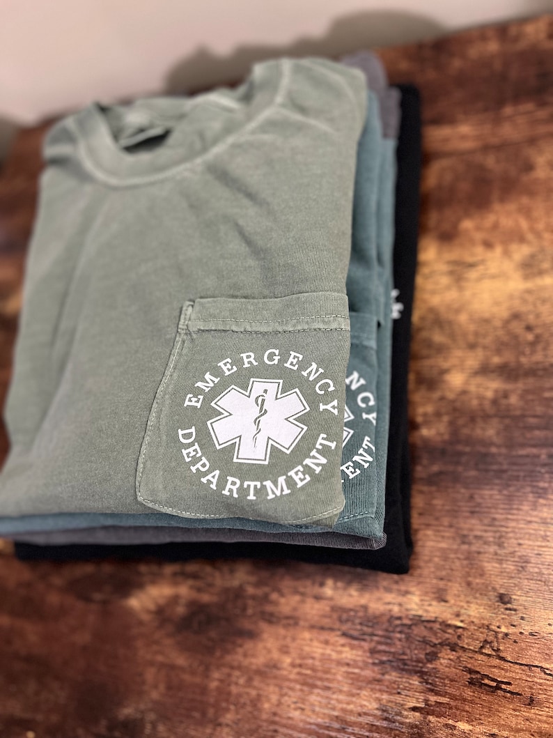 May include: A stack of folded t-shirts in shades of green, gray, and black. The top shirt is olive green with a pocket featuring the words "Emergency Department" around a white medical symbol. The shirts are neatly arranged on a wooden surface.