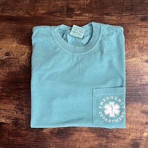 May include: A light teal pocket t-shirt with the words "Emergency Department" and a medical symbol printed on the pocket. The shirt is folded and lies on a dark wood surface.