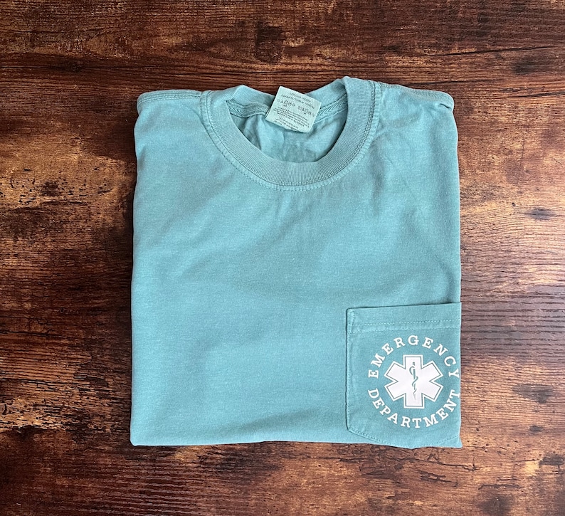 May include: A light teal pocket t-shirt with the words "Emergency Department" and a medical symbol printed on the pocket. The shirt is folded and lies on a dark wood surface.