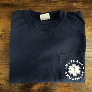 May include: Navy blue pocket t-shirt with the words "Emergency Department" encircling a white medical symbol. The shirt is folded neatly on a wooden surface.