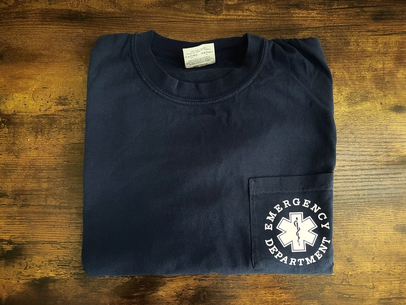 May include: Navy blue pocket t-shirt with the words "Emergency Department" encircling a white medical symbol. The shirt is folded neatly on a wooden surface.