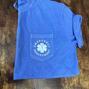 May include: A blue t-shirt with a pocket featuring the text "EMERGENCY DEPARTMENT" in white. The pocket design includes a white medical symbol. The shirt is folded on a wooden surface.